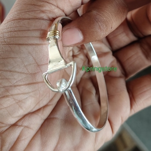 May include: A silver-colored bracelet with a unique clasp design. The clasp features a rectangular plate with a D-shaped loop and a gold-colored accent. The bracelet is held in a hand, showcasing its polished finish and intricate details.