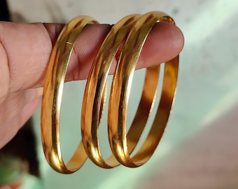 May include: Three gold bangles are held between fingers. The bangles are smooth, shiny, and a warm gold color. The light reflects off the curved surfaces, creating highlights and shadows. The bangles are simple and elegant.