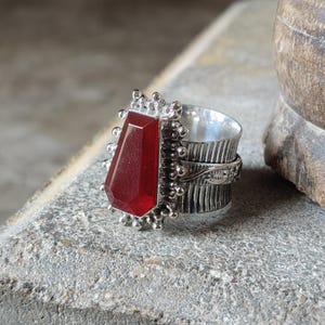 May include: A silver ring with a red gemstone set in a textured silver band. The ring is adorned with small silver beads around the gemstone.
