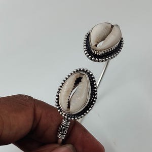 Op de afbeelding: Een open armband met twee witte kaurischelpen als middelpunt. Elke schelp is gezet in een donker, geoxideerd zilverkleurig metalen frame met een kralenrand. De band is gemaakt van een gladde, gepolijste zilverkleurige metalen draad.