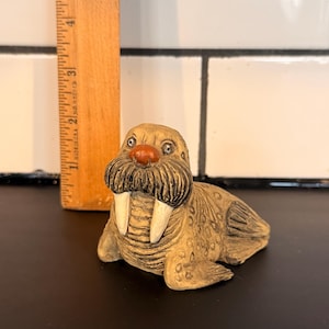 May include: A tan walrus figurine with a brown mustache and white tusks. The walrus has a red nose and is lying down. A wooden ruler is next to the walrus, showing measurements in inches.
