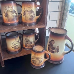 May include: A collection of vintage ceramic beer steins and a pitcher. The steins and pitcher feature sepia-toned illustrations of monks. The steins are displayed on a wooden shelf. The pitcher has a large handle and a wide base.