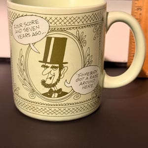 May include: A light green ceramic mug with a cartoon illustration of Abraham Lincoln in a top hat. The mug features text bubbles with the phrases "FOUR SCORE AND SEVEN YEARS AGO..." and "SOMEBODY GOT A RAISE AROUND HERE!"