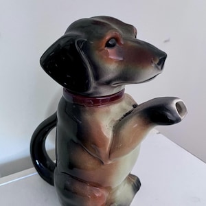 Puede incluir: Una tetera de cerámica con forma de perro salchicha en posición sentada. La tetera con forma de perro presenta una cabeza y orejas negras, un cuerpo marrón y canela y un collar rojo. El pico está situado en la pata delantera del perro. El asa es una forma negra curvada.