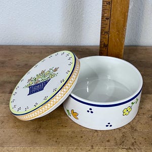 May include: White ceramic round trinket box with a lid. The lid features a blue basket of flowers design. The box has a blue rim and is decorated with yellow flowers and blue dots. The box is open, revealing the interior.