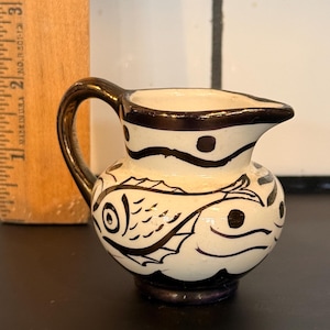 May include: Small, white ceramic creamer with black fish and dot designs. The handle and rim are dark brown. The creamer has a small spout for pouring. A wooden ruler is in the background.