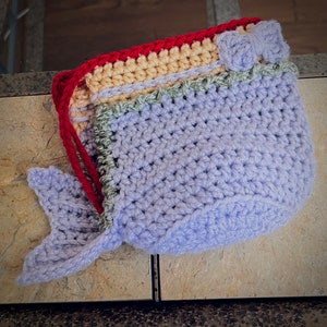 May include: A handmade crochet bag in the shape of a whale. The bag is primarily light purple with a red drawstring and a small bow. The whale's tail is also light purple. The top edge has layers of red, tan, and green.