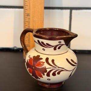 May include: A small, cream-colored ceramic pitcher with a brown handle and rim. The pitcher is decorated with a floral design in shades of burgundy and red. A wooden ruler is placed behind the pitcher.