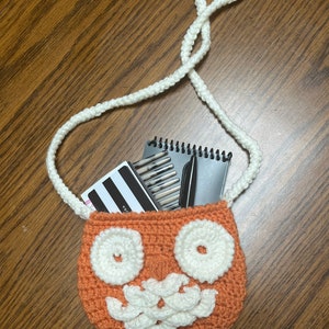 May include: A handmade orange crochet bag with a white face design, including two circular eyes and a mouth. The bag has a white strap and is filled with notebooks and pens. The bag is sitting on a wooden surface.