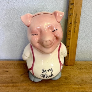 May include: A ceramic pig figurine with a smiling face, closed eyes, and the text "For my Mink" on its belly. The pig is pink with a white shirt and gray pants. A wooden ruler is in the background.