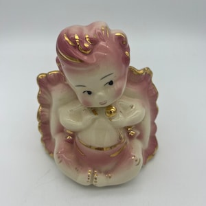 May include: A vintage ceramic planter shaped like a baby girl sitting on a pillow. The planter is painted in shades of pink and white with gold accents.