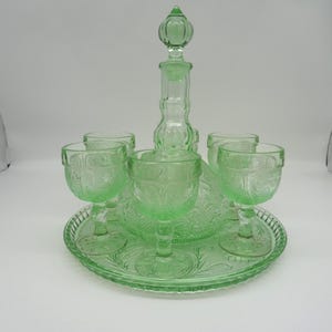 May include: A vintage green glass drink set, including a decanter with a stopper, six stemmed glasses, and a round tray. The set features an embossed floral design. The glassware is a light green color.