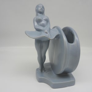 May include: A light blue ceramic figurine of a woman with a skirt-like form, holding a vase. The figurine is standing on a rectangular base. The vase is a curved, open shape. The overall design is a stylized, art deco aesthetic.