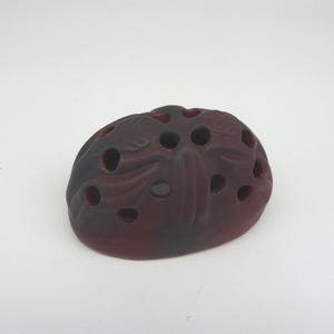 May include: A dark red, organic-shaped decorative object with multiple circular holes. The object has a textured surface and appears to be made of ceramic or a similar material. The color transitions from a deep red to a darker, almost black hue.