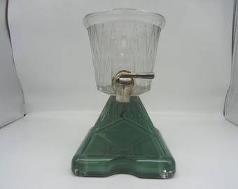 May include: Vintage clear glass beverage dispenser with a diamond pattern and a silver spigot. The dispenser sits on a green, geometric base. A classic piece for serving drinks at gatherings.