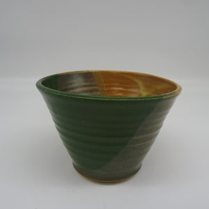May include: A handmade ceramic bowl with a green exterior and a brown interior. The bowl has a wide, open top and a slightly tapered shape. The exterior features horizontal ridges, adding texture. A unique piece for serving or display.