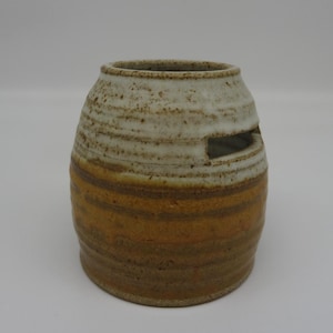 May include: A handmade ceramic vase with a cylindrical shape and a horizontal rectangular opening. The vase features a layered design with bands of white, brown, and orange hues. The pottery has a rustic, textured appearance.