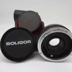 May include: A black Soligor lens cap with white text is in front of a camera lens. The lens has a silver ring and a black body. A black camera case is in the background.