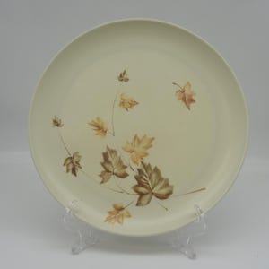 May include: A cream-colored, round dinner plate with a leaf design. The leaves are in shades of brown and tan, with delicate stems. The plate is displayed on a clear plastic stand, showcasing its decorative pattern.