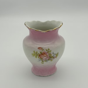 May include: A small, pink porcelain vase with a scalloped rim and a floral design featuring a rose. The vase has a gold trim around the rim.