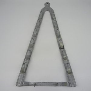 Puede incluir: Un objeto metálico triangular de color plateado con un lazo en la parte superior y una barra horizontal en la parte inferior. Los dos lados angulados presentan una serie de recortes rectangulares. El objeto parece ser una herramienta o un componente de hardware vintage.