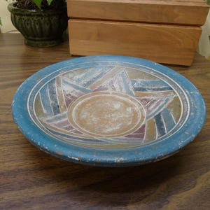 May include: A shallow circular ceramic bowl with a wide, textured blue rim. The interior features a central tan circle surrounded by a geometric pattern of alternating triangles in shades of muted blue, maroon, and tan, separated by thin white lines.