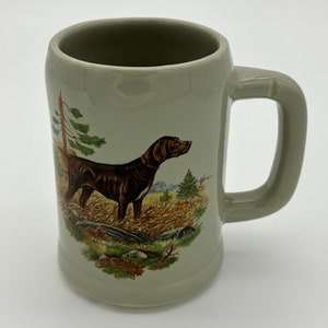 May include: A ceramic mug with a brown and white hunting dog illustration. The dog is standing in a field with trees and bushes in the background. The mug is a light green color.