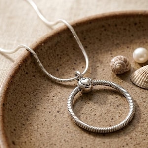May include: A silver necklace featuring a circular pendant with a heart-shaped charm. The necklace is presented on a brown plate, alongside seashells and a pearl. The pendant has a textured, rope-like design.