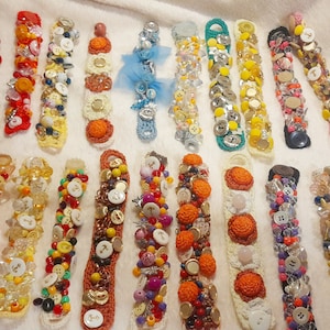 Puede incluir: Una colección de 20 pulseras coloridas hechas con botones, cuentas y tejido de crochet. Las pulseras son de diferentes colores y patrones, y están dispuestas en una fila sobre una superficie blanca.