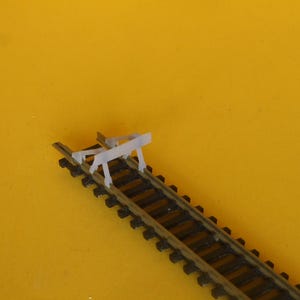 May include: A miniature model train track with a light gray barrier. The track is black and brown, set against a yellow background. The barrier is positioned on the track, suggesting a stop or safety feature for model train enthusiasts.