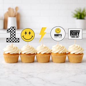 May include: Five cupcakes topped with decorative paper toppers: a black and white checkered number one, a yellow smiley face with lightning bolt eyes, a yellow lightning bolt, a circular sign reading RORY'S FIRST TOUR, and a rectangular sign reading RORY TURNS ONE.