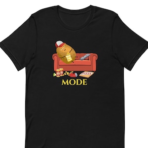 Könnte beinhalten: Schwarzes T-Shirt mit einer Cartoon-Kartoffel, die eine Baseballkappe trägt und auf einem roten Sofa mit Fernbedienung, Chips, Pizza und Limonade entspannt. Der Text "MODE" befindet sich unter dem Sofa.