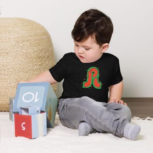 May include: A black toddler t-shirt featuring a large red letter "R" outlined in green, decorated with small white lights. The child is wearing gray pants and sitting on a white surface, playing with colorful blocks.