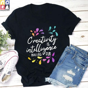May include: Black t-shirt with a colorful design and the text "Creativity is intelligence having fun".