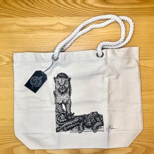 May include: A white canvas tote bag with rope handles. The bag features a black and white illustration of a leopard and a panther cub. The text "leopard (panthera pardus)" is printed below the illustration.