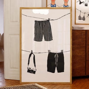 Peut inclure: Une affiche encadrée avec des illustrations en noir et blanc de vêtements et d'un appareil photo sur une corde à linge. L'affiche présente un short rayé, un short uni et un appareil photo. Deux autres affiches encadrées sont visibles en arrière-plan.