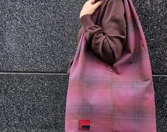 May include: A large tote bag with a plaid pattern in shades of pink, purple, and gray. The bag has a rectangular red patch with black detailing. The bag is made of a soft fabric and has a button closure.