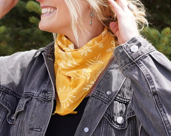 Op de afbeelding: Een vrouw draagt een bordeauxrode baseballpet, een gele bandana met een witte zon en een bloemenpatroon, en een grijs denimjack. Ze glimlacht en kijkt naar rechts.