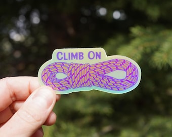 Op de afbeelding: Een holografische sticker met de tekst "CLIMB ON" boven een paars en bruin klimtouwontwerp. De sticker wordt tegen een vage groene achtergrond gehouden.