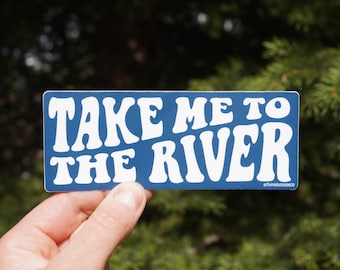 Op de afbeelding: Een rechthoekige blauwe sticker met de witte tekst "TAKE ME TO THE RIVER", gehouden tegen een achtergrond van groen gebladerte. De sticker heeft afgeronde hoeken en een retro lettertype.