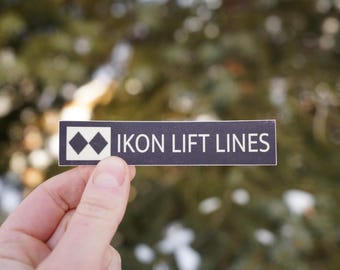 Op de afbeelding: Een zwart-witte sticker met de tekst "IKON LIFT LINES" en een wit bergpictogram met twee zwarte diamanten.