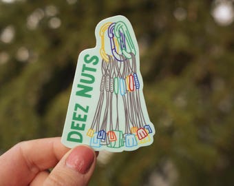 Op de afbeelding: Een groene en witte sticker met de tekst "DEEZ NUTS" en een kleurrijke illustratie van een groep klimmers.