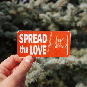 May include: An orange sticker with the words "SPREAD the LOVE" in white, along with a skier graphic. The sticker is held in front of a blurred green and blue background.