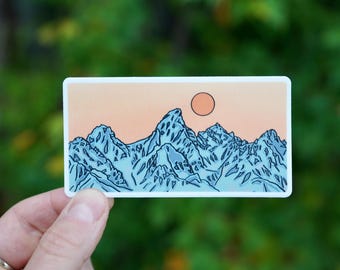 Op de afbeelding: Een sticker met een blauwe en witte illustratie van een bergketen met een enkele oranje zon die achter de toppen ondergaat.