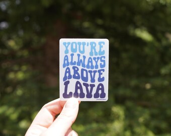 Op de afbeelding: Een rechthoekige sticker met de tekst "YOU'RE ALWAYS ABOVE LAVA" in blauwtinten. De sticker wordt tegen een vage groene achtergrond gehouden.