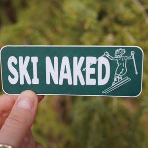 May include: A rectangular sticker with rounded corners, featuring the words "SKI NAKED" in bold white letters on a dark green background. A skier graphic is on the right. The sticker is held up against a blurred green background.