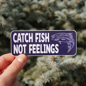 Puede incluir: Una pegatina rectangular con esquinas redondeadas, con el texto "CATCH FISH NOT FEELINGS" en letras mayúsculas blancas. Un contorno blanco de un pez está a la derecha. La pegatina se sostiene frente a un fondo verde borroso.