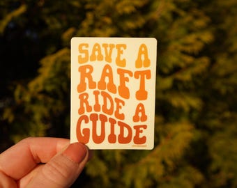 Op de afbeelding: Een rechthoekige sticker met de tekst "SAVE A RAFT RIDE A GUIDE" in oranje en gele retro lettertype. De sticker wordt in een hand gehouden tegen een vage groene achtergrond.