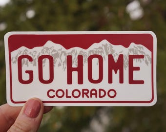 Op de afbeelding: Een witte sticker met een rode rand en een bergketen-ontwerp. De sticker zegt "GO HOME COLORADO" in rode letters.