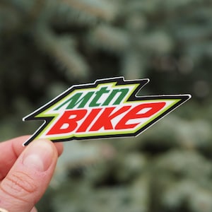 以下が含まれることがあります： 緑と赤で「mtn BIKE」の文字が書かれたステッカー。黒と黄色の縁取りがあり、ギザギザした角度のある形をしています。背景はぼやけた緑色です。